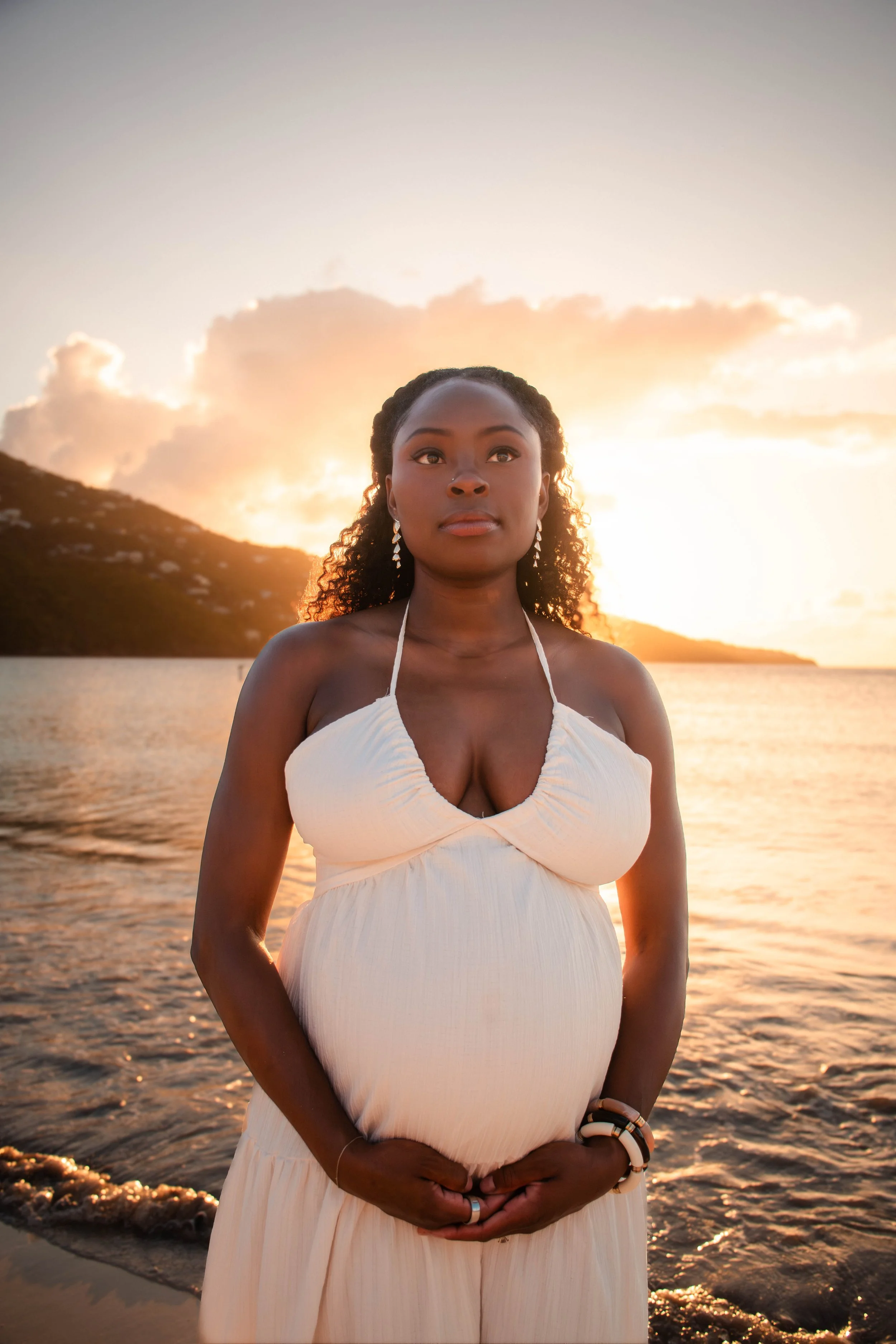 A pregnant woman in a white dress standing on a beach at sunset, hands resting on her belly, with mountains and cloudy sky in the background.