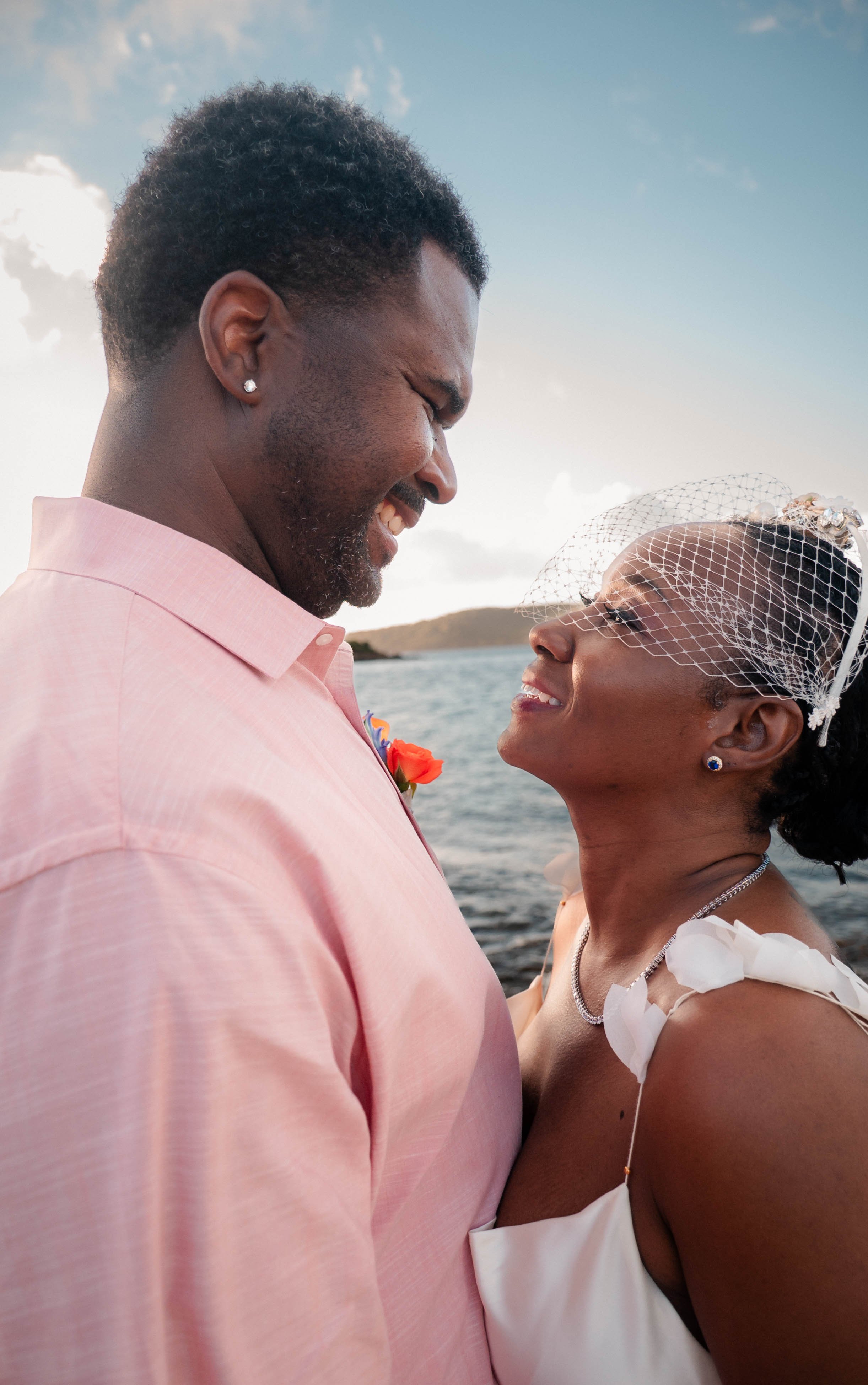 A man and woman looking at each other and smiling outdoors with water and a sky with clouds in the background.