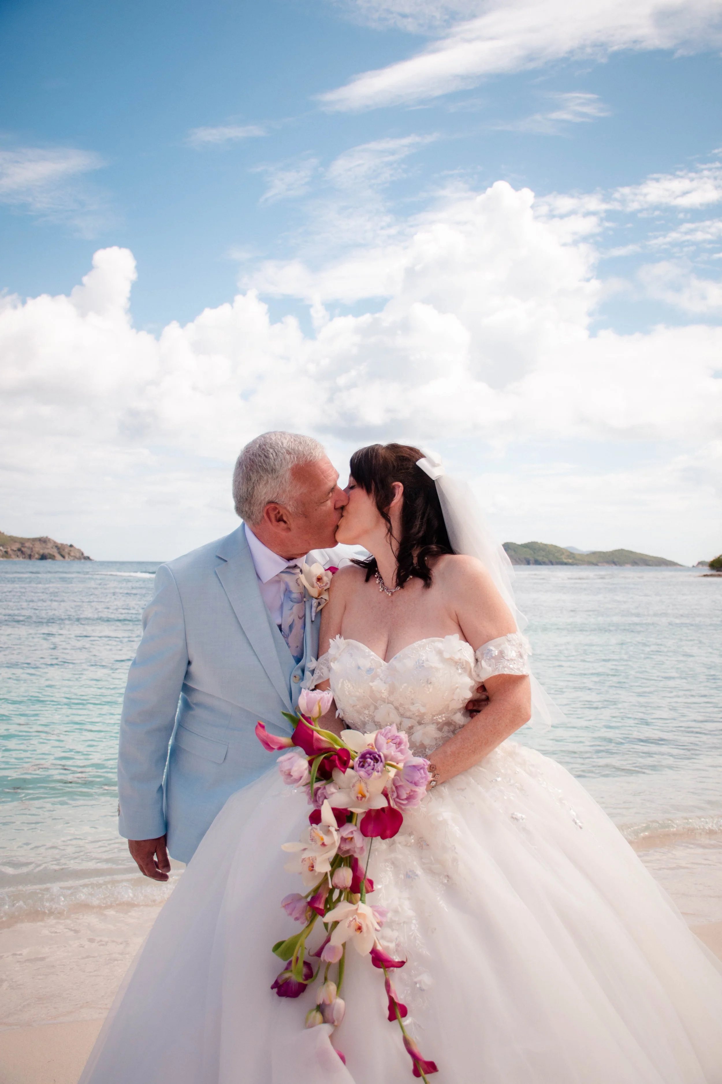 A bride and an older man, likely her father, kissing on a beach with the ocean and cloudy sky in the background. The bride wears a white wedding dress with lace details, holds a cascading bouquet of pink, purple, and white flowers, and has a white ve