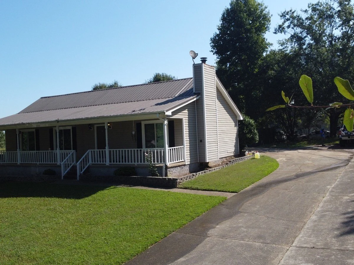 A white house with a porch, grey roof, and chimney, surrounded by green lawn and trees, on a sunny day.