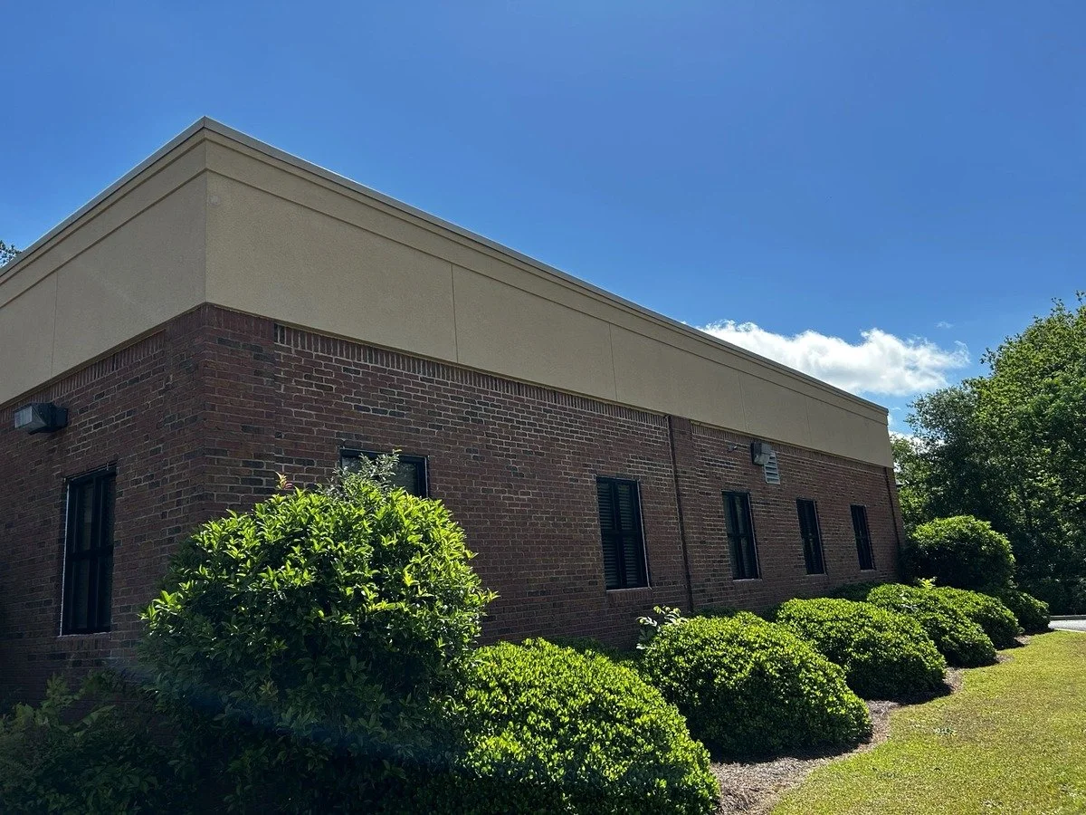 A brick building with beige trim on top, four windows with dark shutters, surrounded by green bushes and trees under a partly cloudy blue sky.