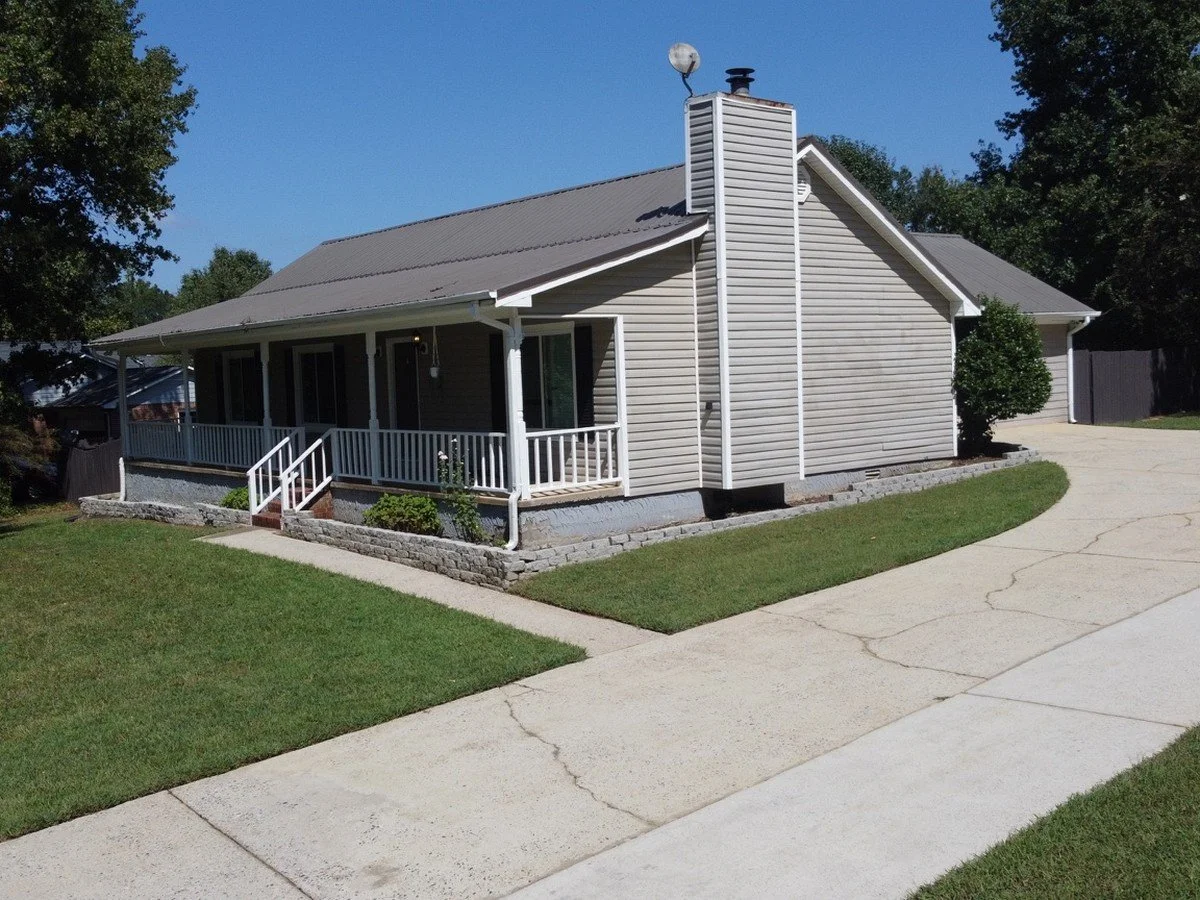 A residential house with beige siding, a gray roof, and a white front porch with railing and steps, surrounded by a manicured lawn and a concrete sidewalk.