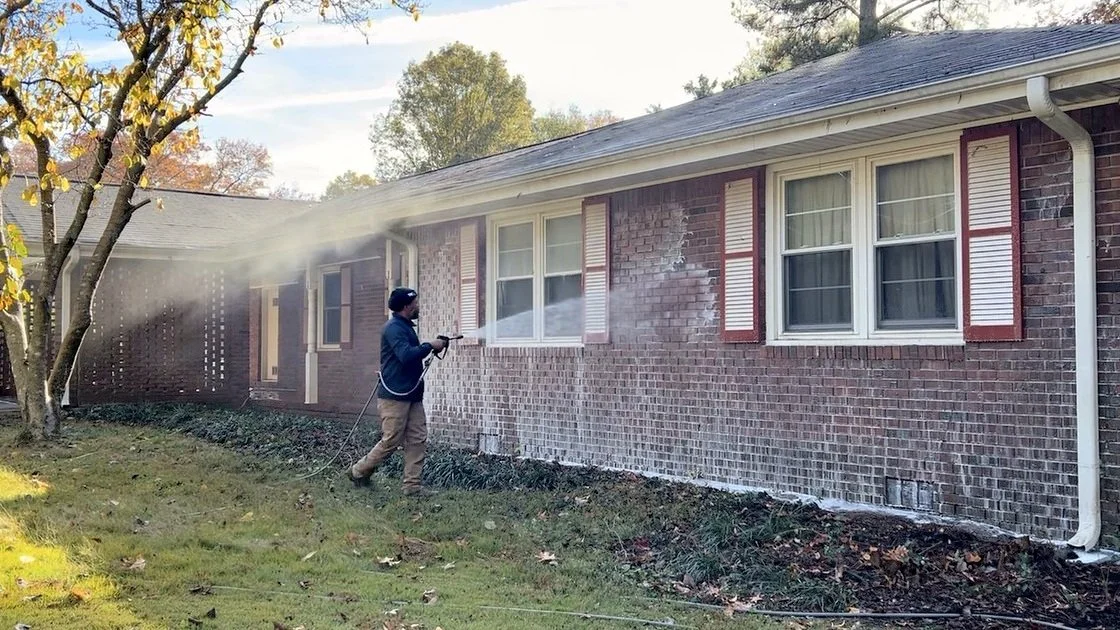 Person pressure washing a brick house exterior with water spray.