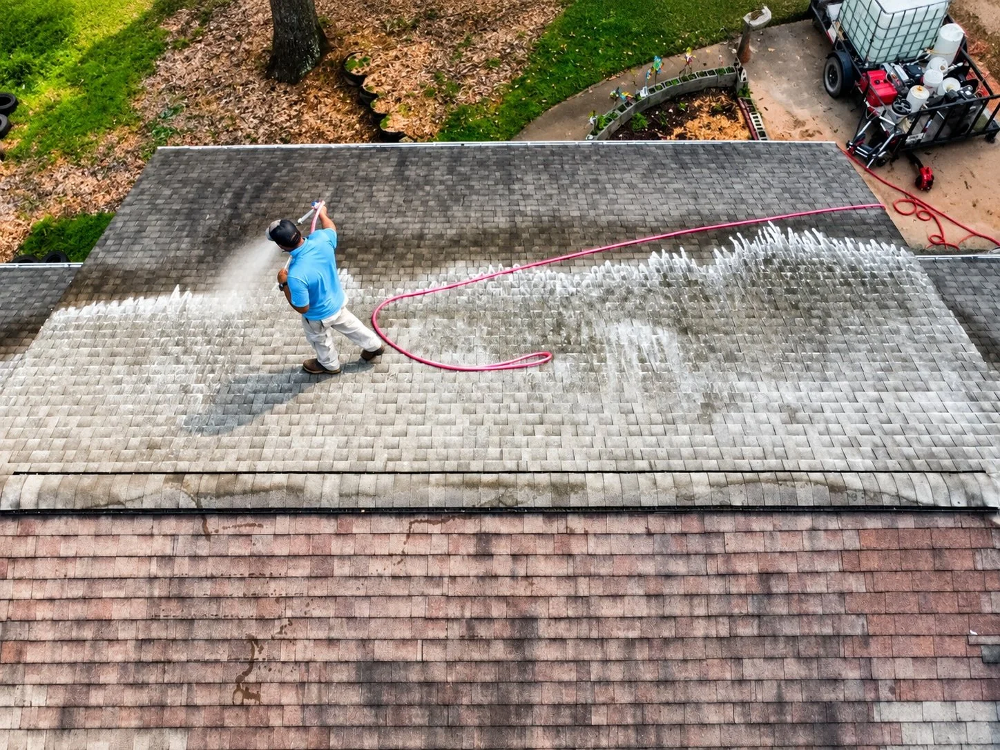 A person wearing a helmet and blue shirt cleaning a residential roof with a pressure washer, causing water and dirt to spray off the shingles.