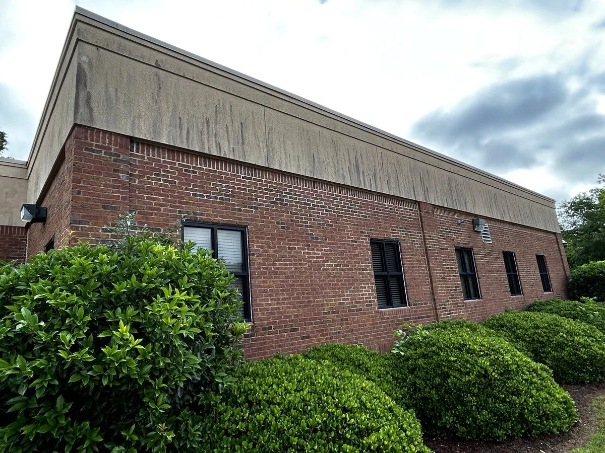 Exterior of a brick building with four windows, surrounded by green bushes, with a cloudy sky overhead.