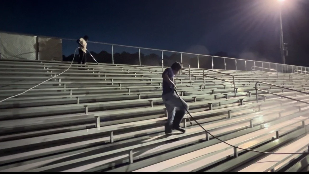 Two people cleaning and washing bleacher seats in an outdoor stadium at night under stadium lights