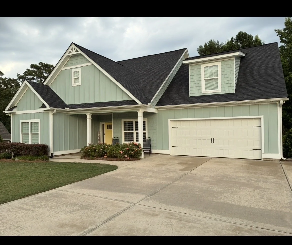 A light green two-story house with a dark gray roof, white trim, a front porch with two chairs, a welcoming sign, and a closed garage door. The house has small dormer windows and is surrounded by a manicured lawn with bushes and pink flowers.