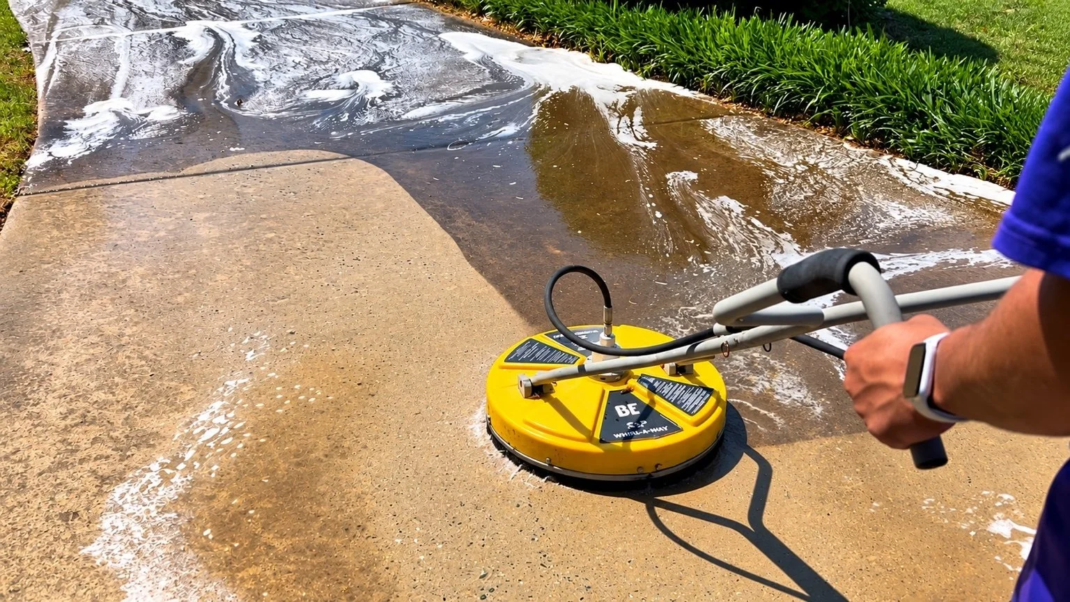 A person using a yellow pressure washer to clean a concrete sidewalk, with soap and water visible on the surface.