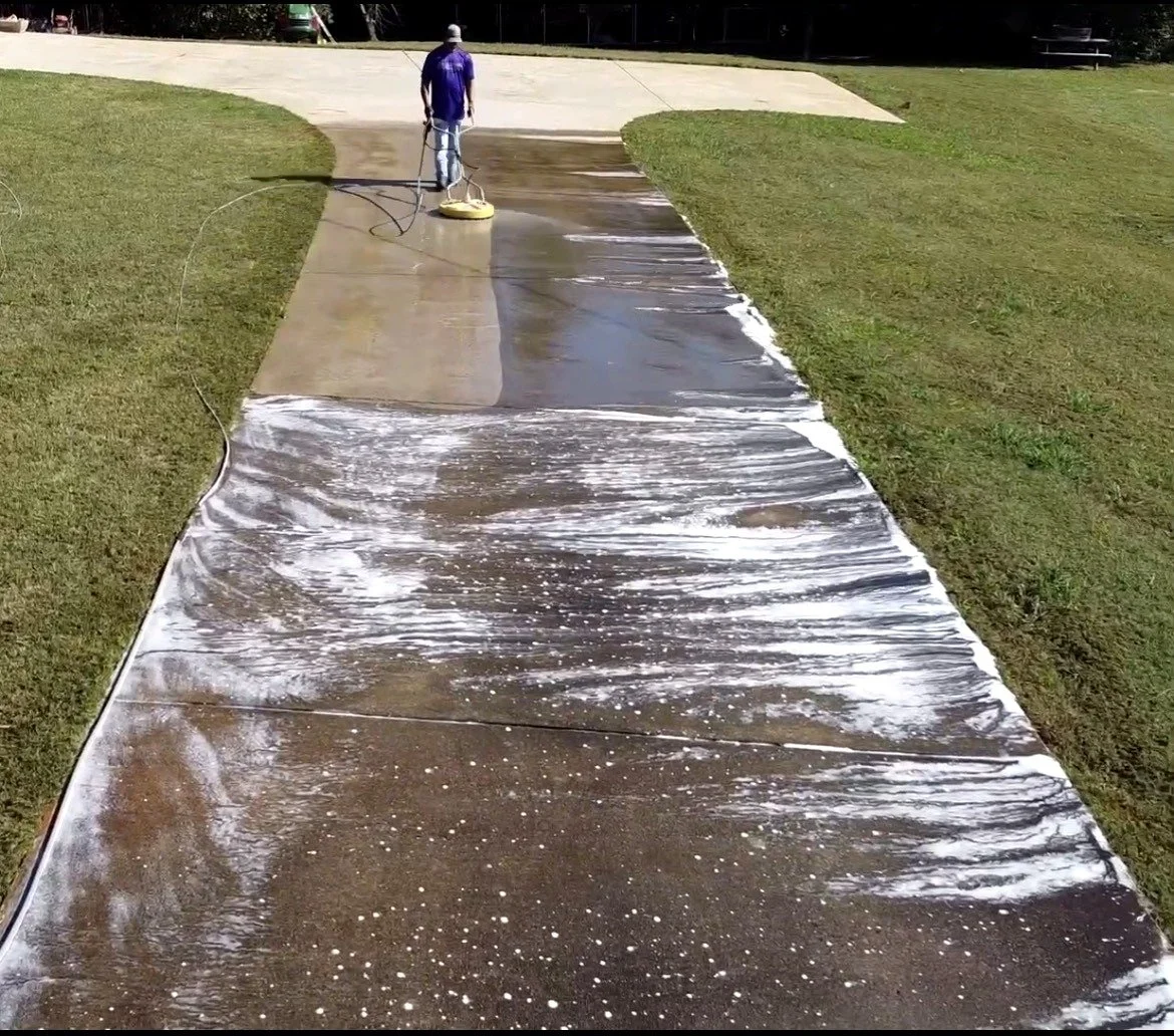 Person pressure washing a concrete sidewalk, with soap and water, outdoors on a sunny day.