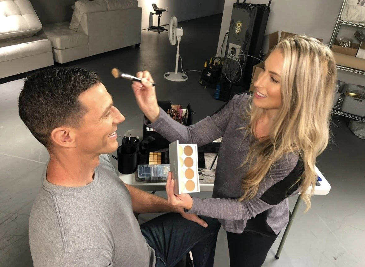 A makeup artist applying makeup on a smiling man sitting in front of her in a room with sofas, a fan, and various makeup supplies.
