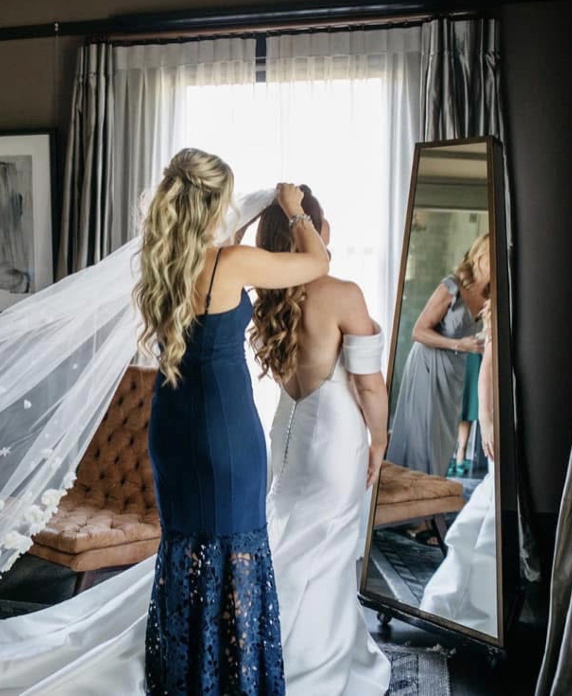 A bride in a white wedding dress is being helped with her hair by a woman in a blue dress. They are standing in front of a mirror in a room with large windows and curtains. The bride is holding a white arm sleeve, and the woman in blue is styling her hair.