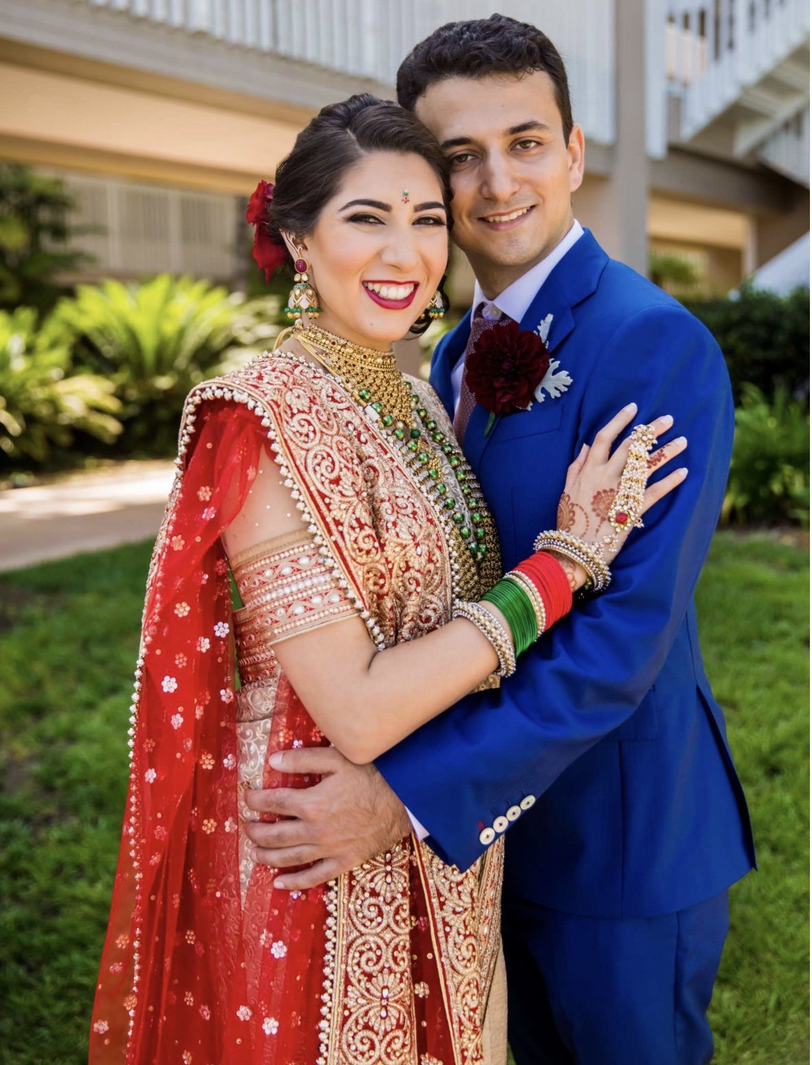 A couple dressed in traditional Indian wedding attire, smiling and embracing outdoors, with greenery and a building in the background.