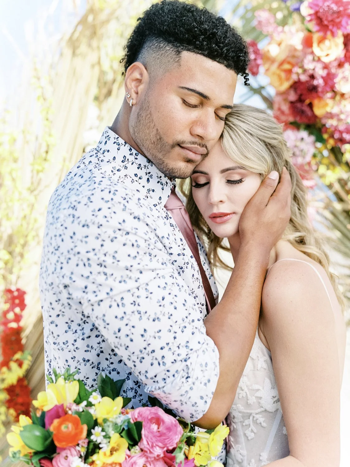 A couple embracing, with the man gently holding the woman's face, standing outdoors with flowers in the background and foreground.