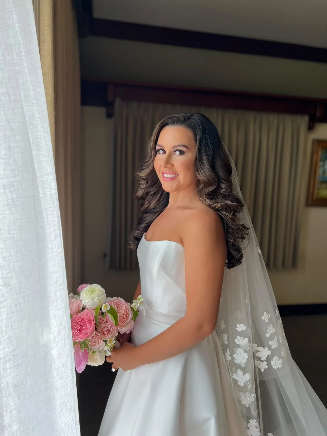 A bride in a white wedding dress holding a bouquet of pink and white flowers, smiling and looking at the camera indoors near a window with curtains.