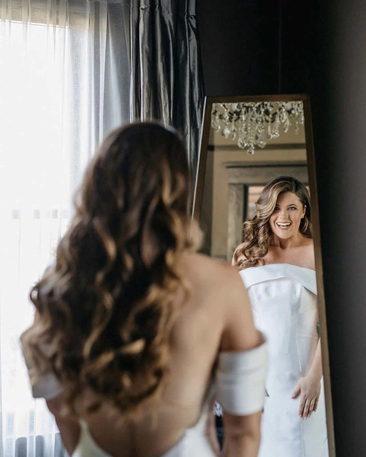A woman with long, wavy hair wearing a white dress smiles at her reflection in a full-length mirror in a room with dark walls, a chandelier, and curtains.