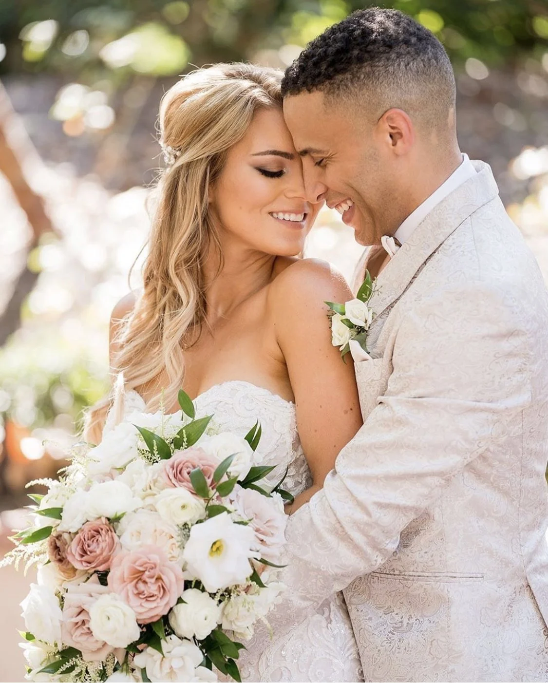 A bride and groom sharing a kiss or intimate moment outdoors, with the bride holding a large bouquet of white and pink flowers, both dressed in wedding attire, smiling with eyes closed.
