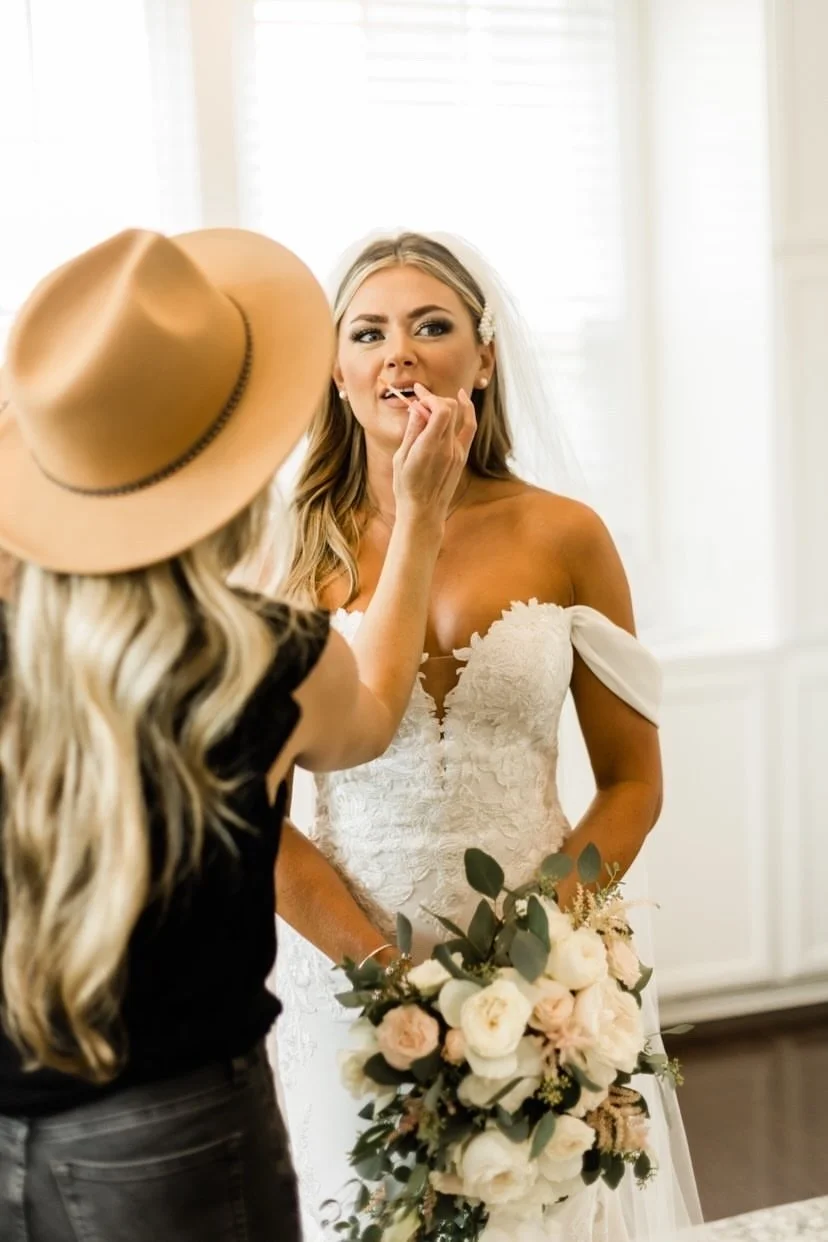 A bride in a white wedding dress holding a bouquet of flowers is getting makeup done by a woman wearing a black outfit and a wide-brimmed hat.