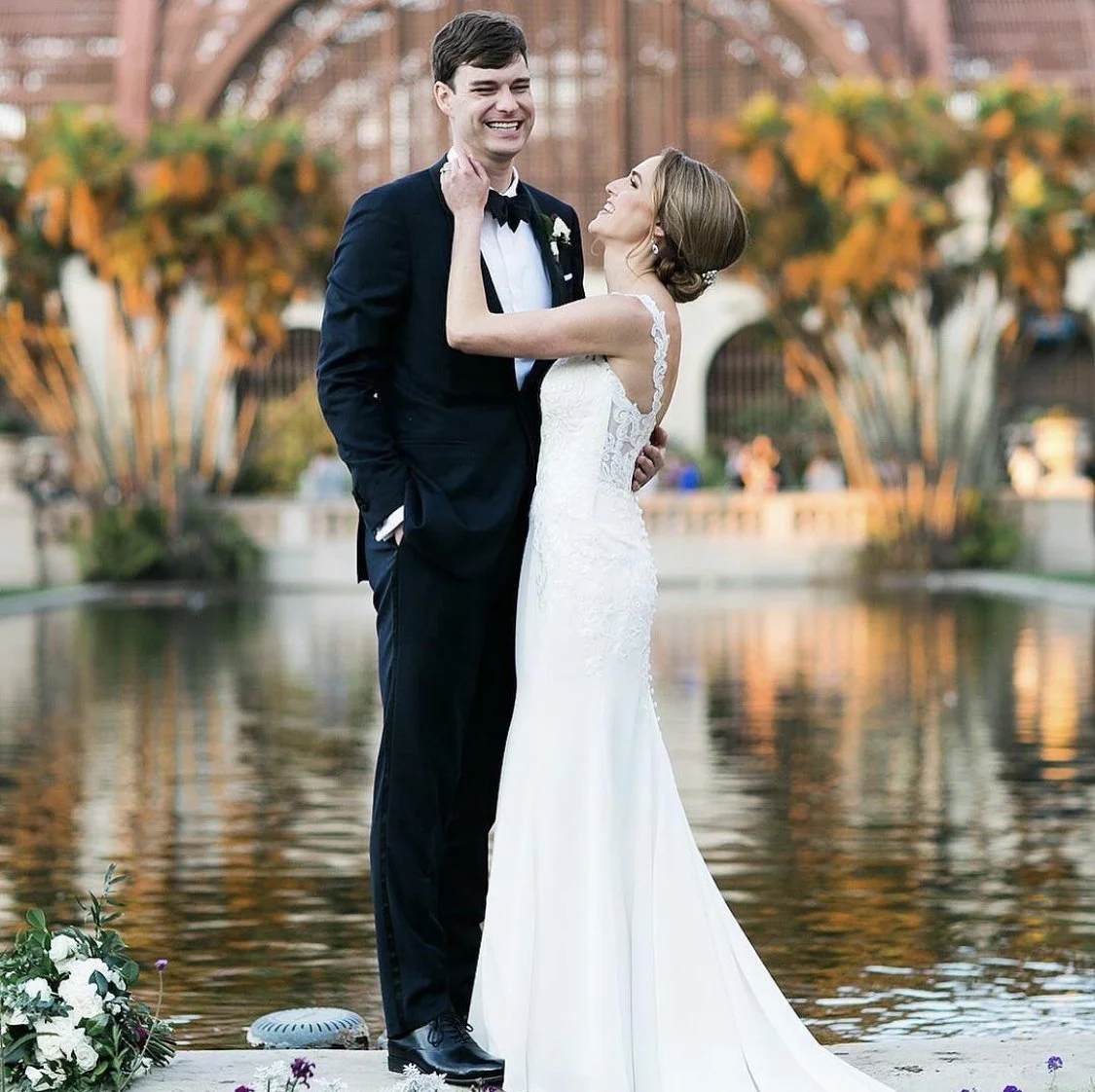 A bride and groom dancing outdoors by a water feature, with autumn trees in the background, during their wedding celebration.