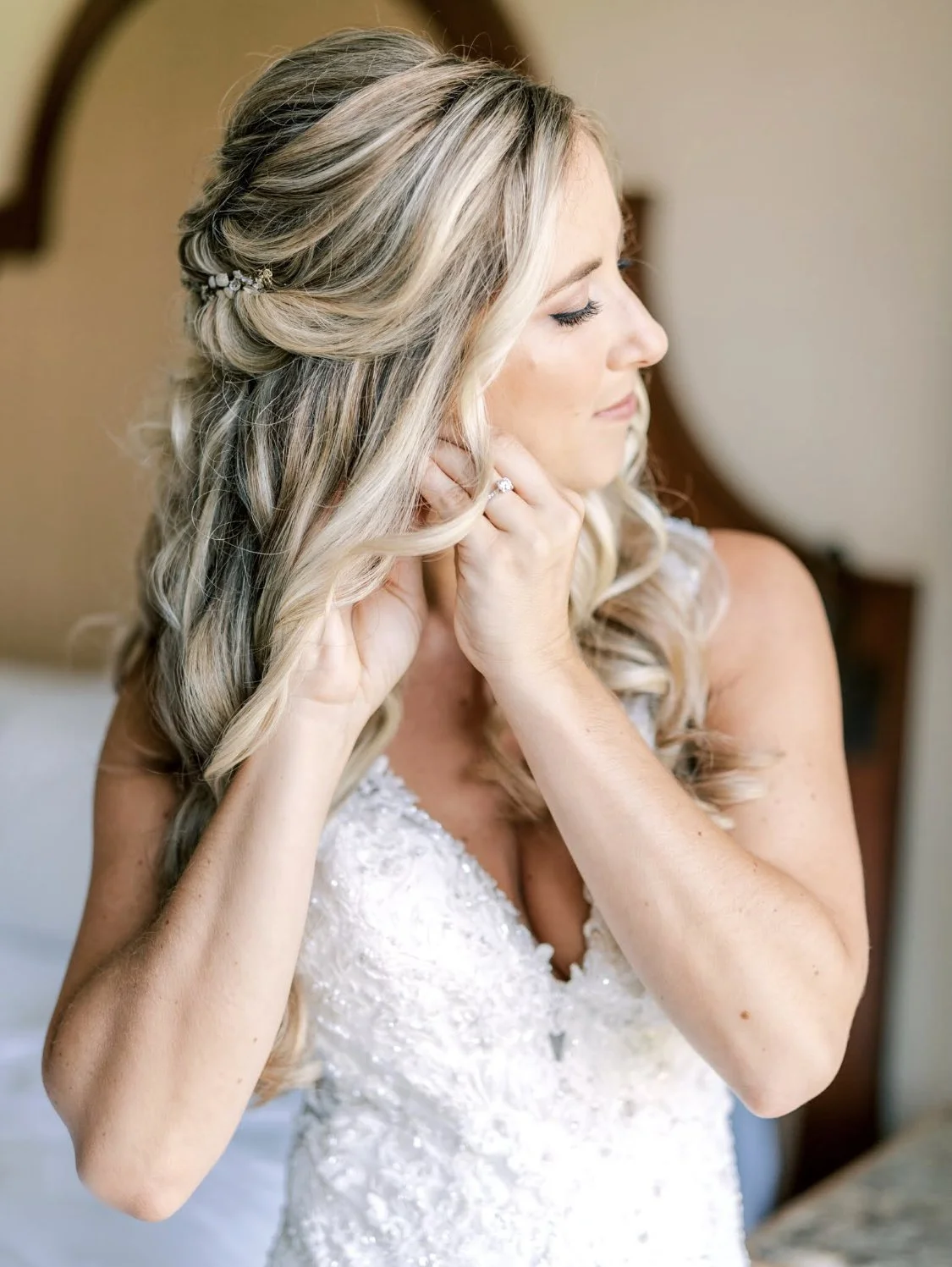 Bridal woman with blonde hair styled in loose waves, wearing a white lace wedding dress, adjusts her earring with one hand, in a warmly lit room.