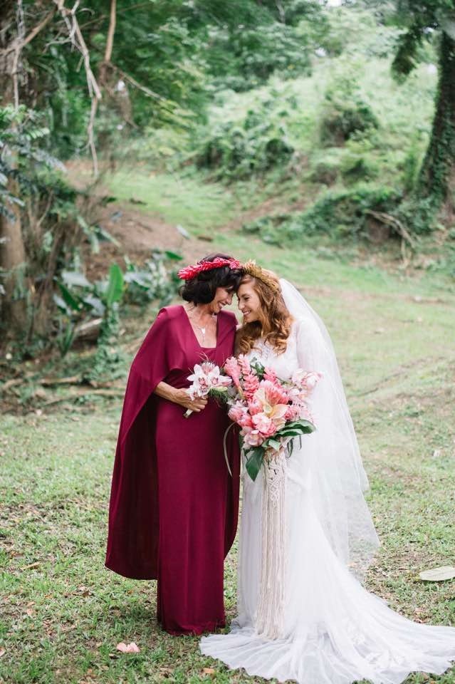 A bride in a white wedding gown holding a bouquet of pink flowers, standing close to a woman in a burgundy dress, both smiling and touching foreheads outdoors in a lush, green forest setting.