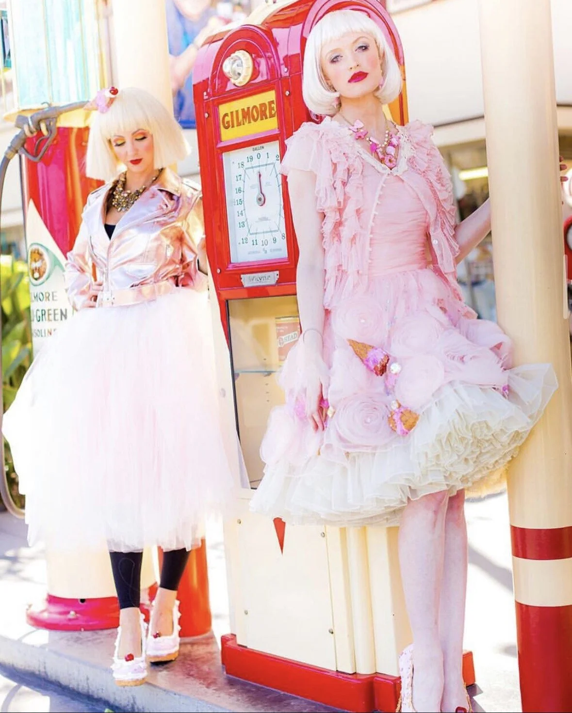 Two mannequins dressed in pastel pink and white clothing, posing in front of a vintage-style red and yellow Gilmore vending machine.