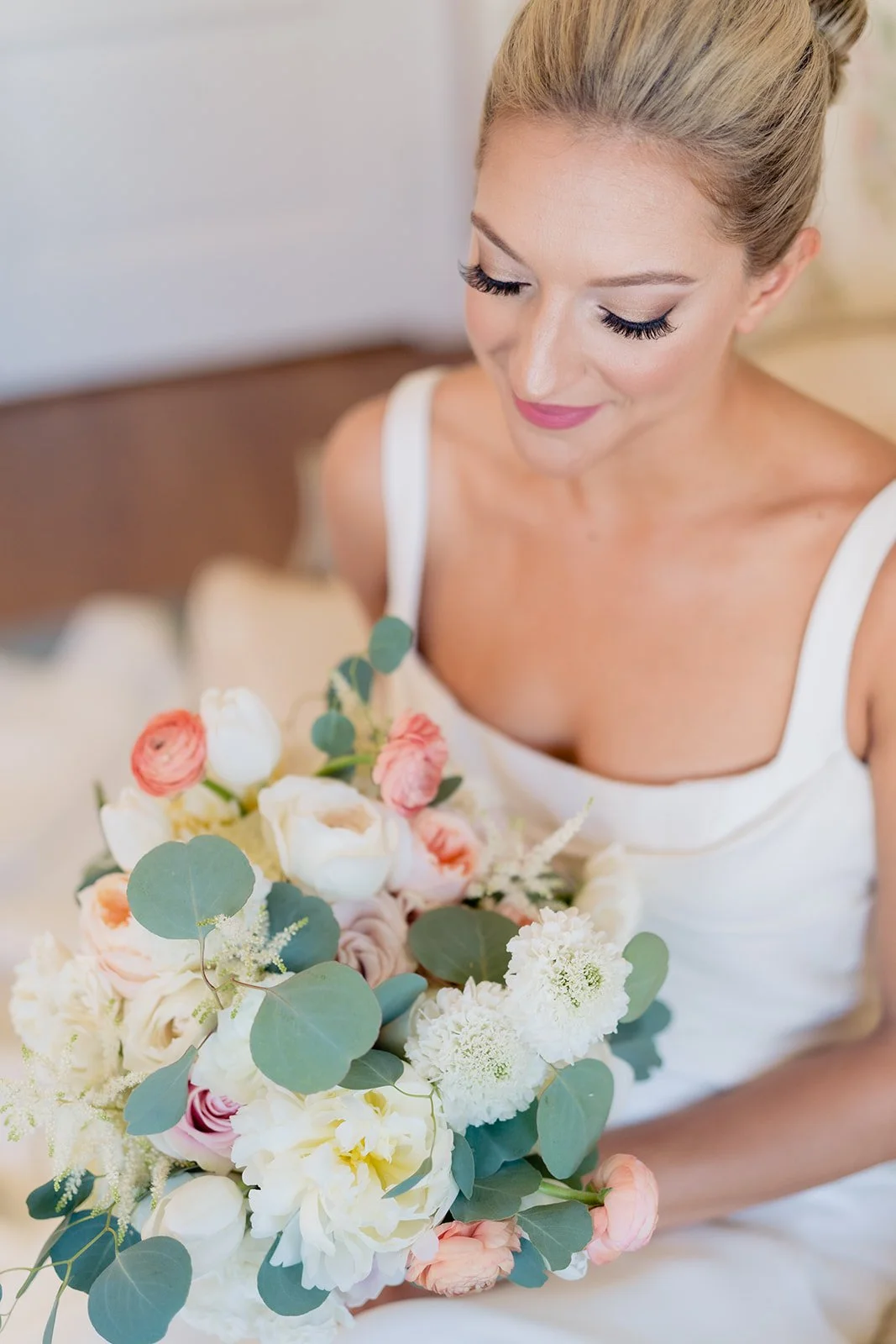 A woman with blonde hair styled in an updo and wearing makeup, holding a large bouquet of white and pink flowers with green leaves, sitting indoors.