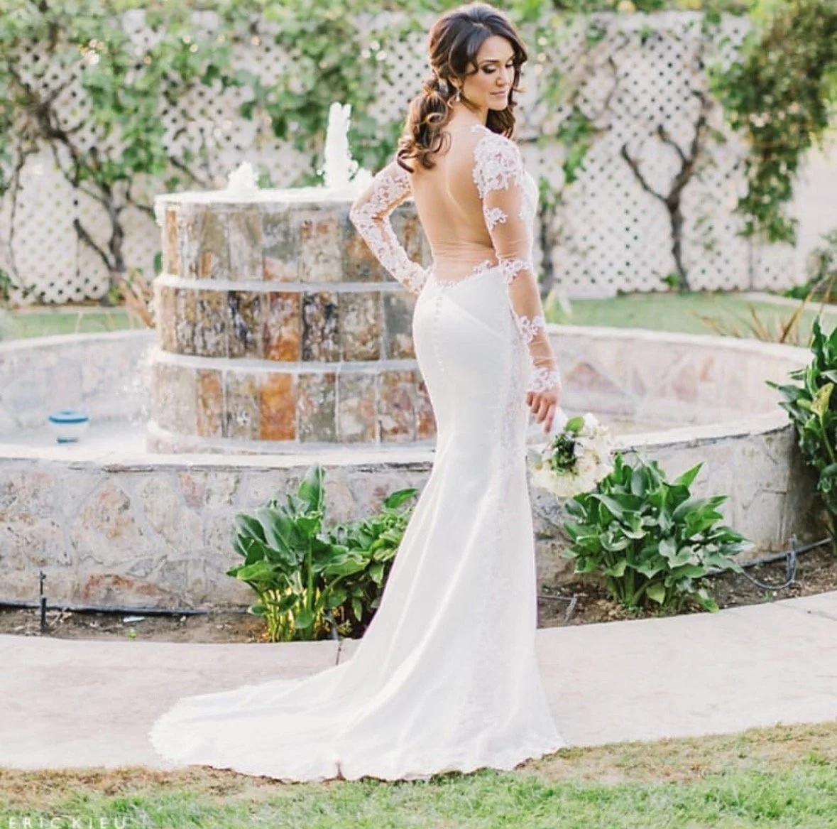 A bride in a white wedding dress with lace sleeves and an open back, holding a bouquet, standing near a stone fountain with greenery in the background.