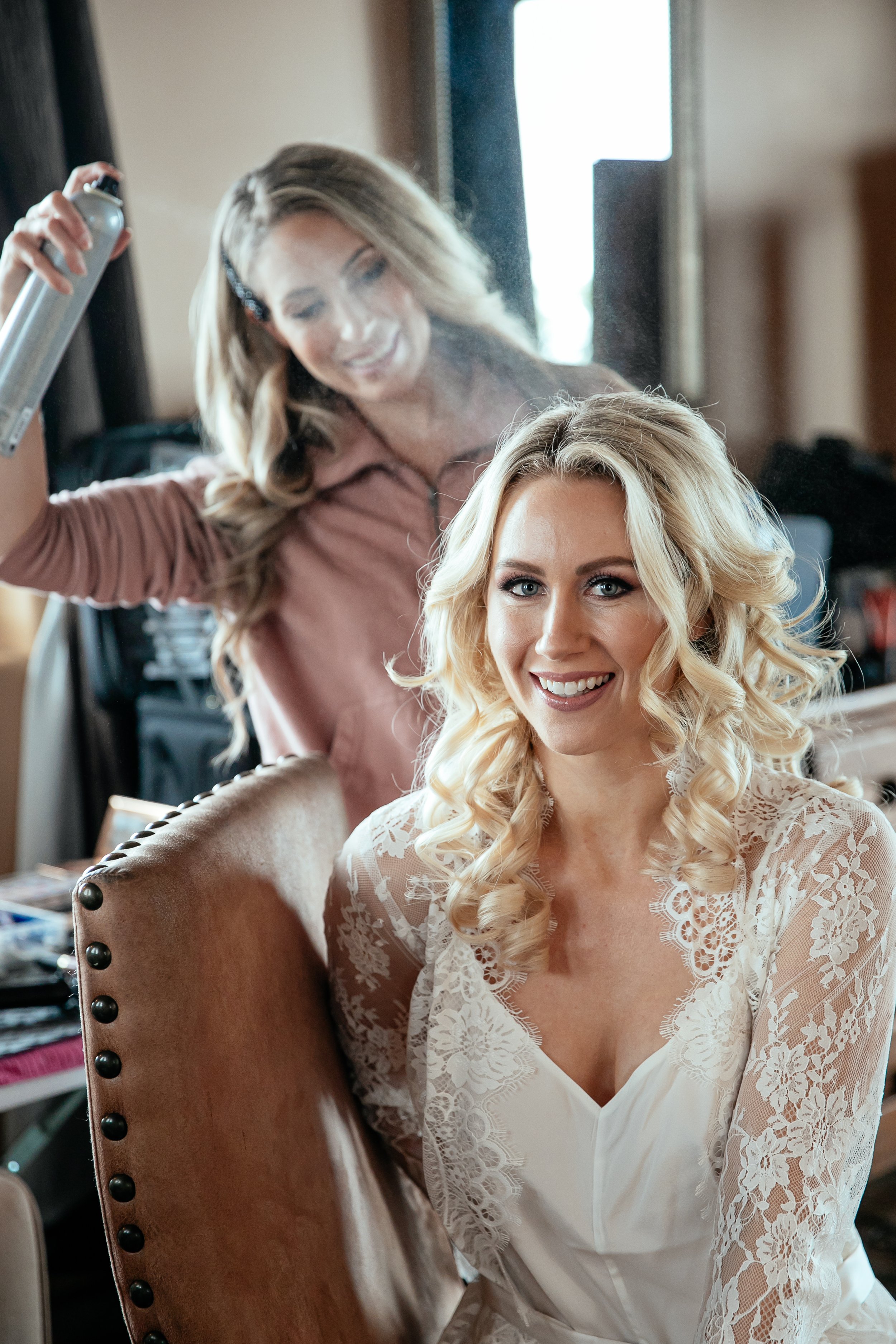 A woman with blonde hair styled in loose curls is sitting in front of a mirror, smiling at the camera, wearing a white lace robe. Another woman with long, light brown hair, wearing a pink top, is standing behind her, styling her hair with a curling iron.