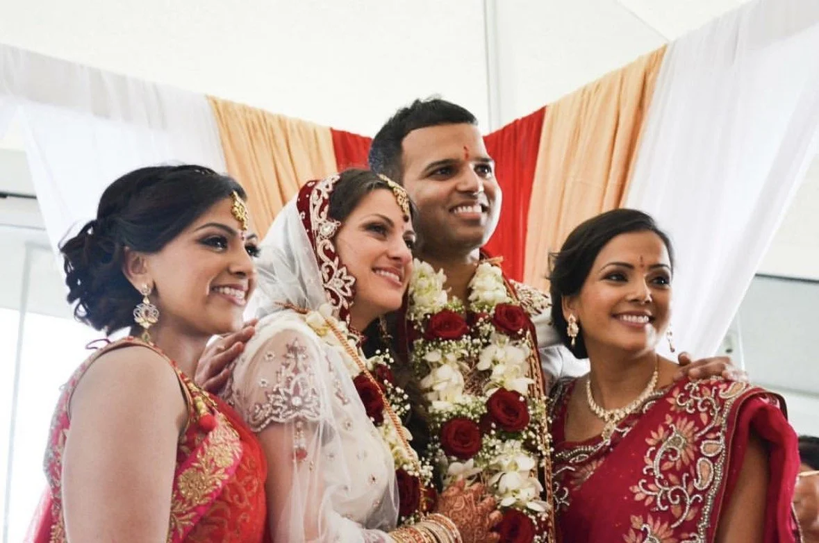 Group of four people dressed in traditional Indian wedding attire at a wedding ceremony, standing in front of a backdrop with white, red, and orange fabric.