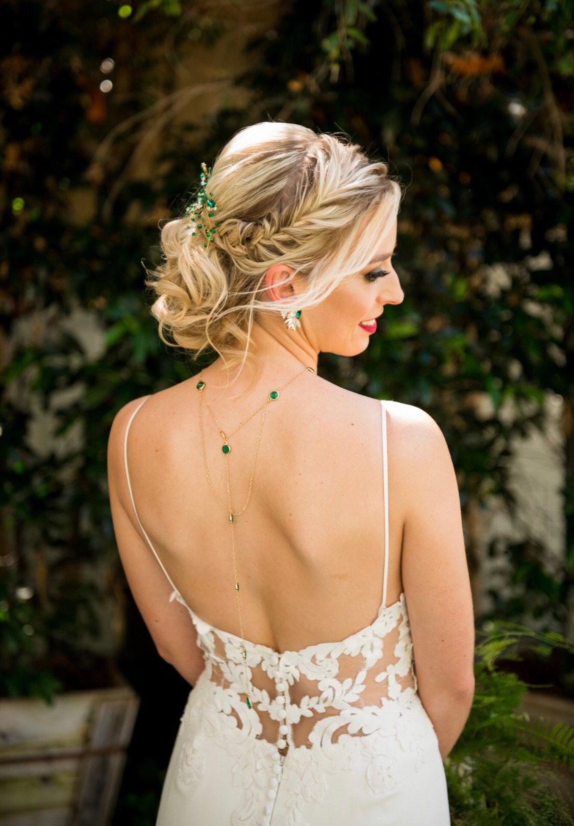 A woman with blonde hair styled in an elegant braid and curls, wearing a white wedding dress with floral lace details, and accessorized with green gemstone jewelry, standing outdoors near greenery.