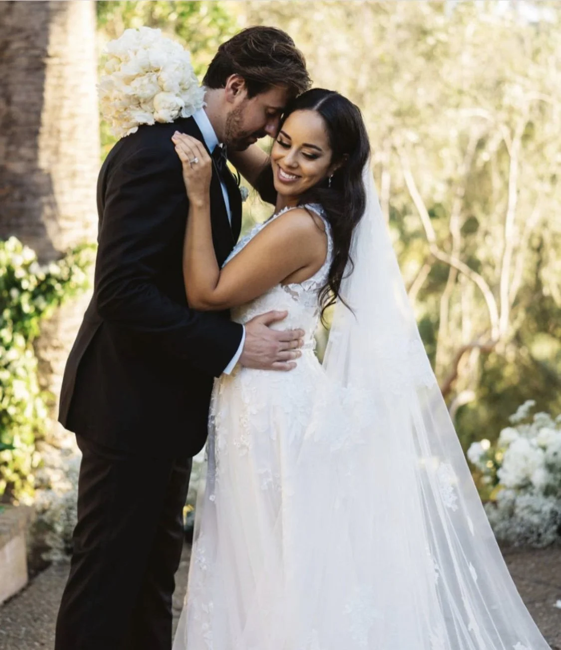 A bride and groom sharing an intimate moment outdoors, with the groom's forehead touching the bride's. The bride is smiling with her eyes closed, wearing a white wedding dress and veil, while the groom is in a black tuxedo holding her waist. There are trees and flowers in the background.