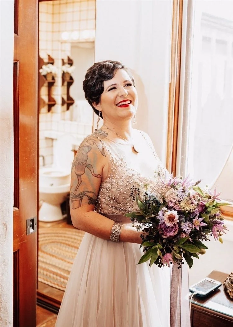 A smiling woman with short dark hair and tattoos on her arms, wearing a beaded light-colored dress, holding a bouquet of flowers, standing near a window in a warmly lit room.