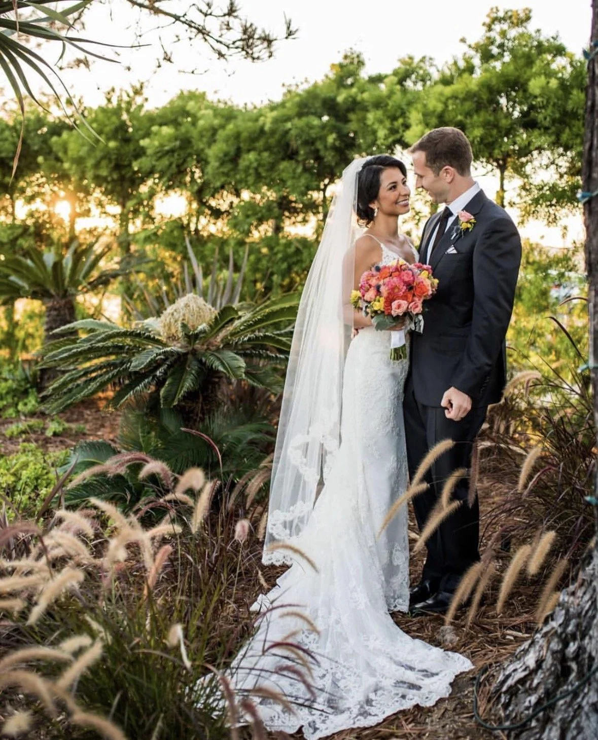 A bride and groom standing close together outdoors during sunset, surrounded by greenery and plants, with the bride holding a colorful bouquet of flowers.