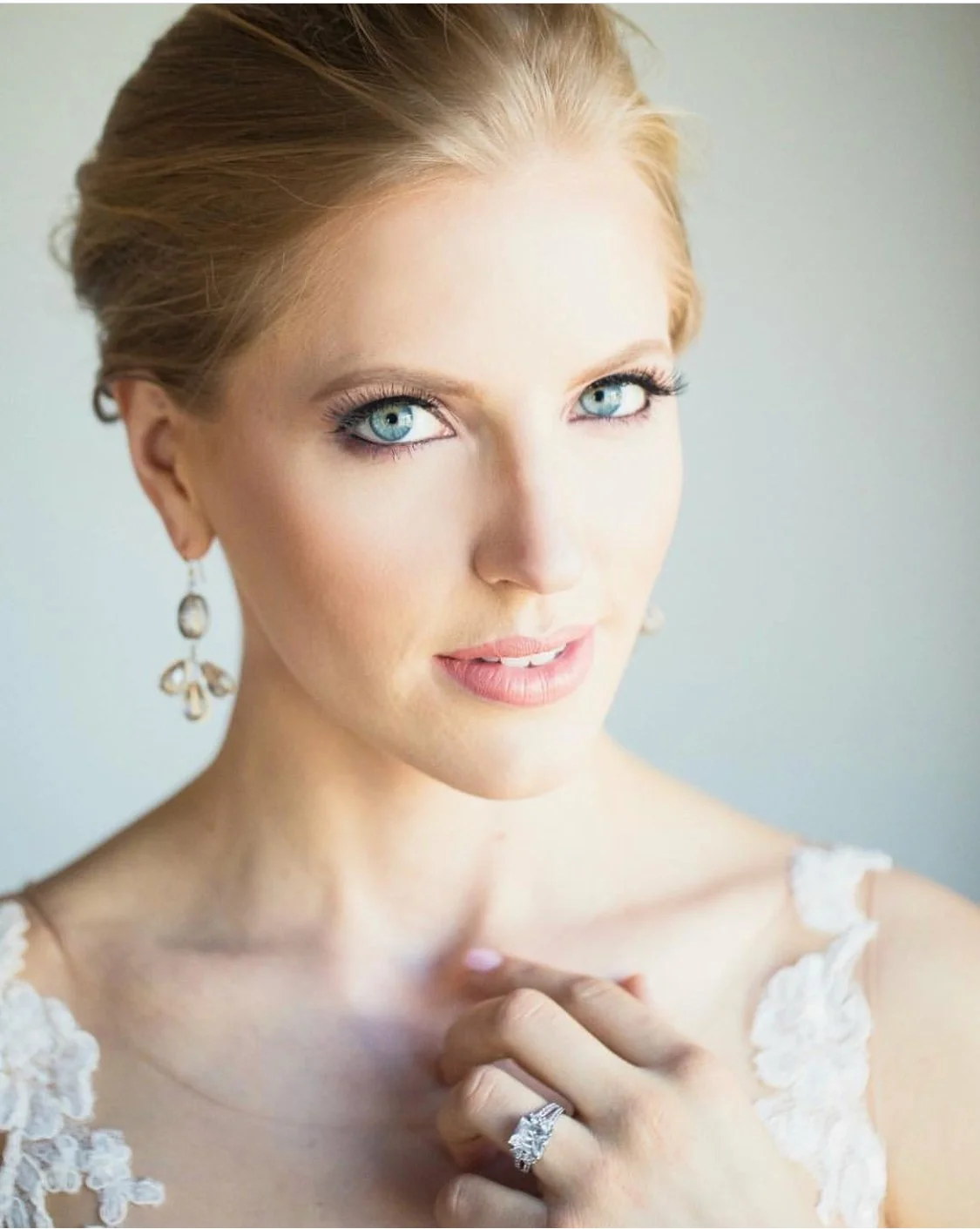 A close-up portrait of a woman with blue eyes and blonde hair, wearing earrings and a ring on her finger, with lace details on her dress, against a light background.