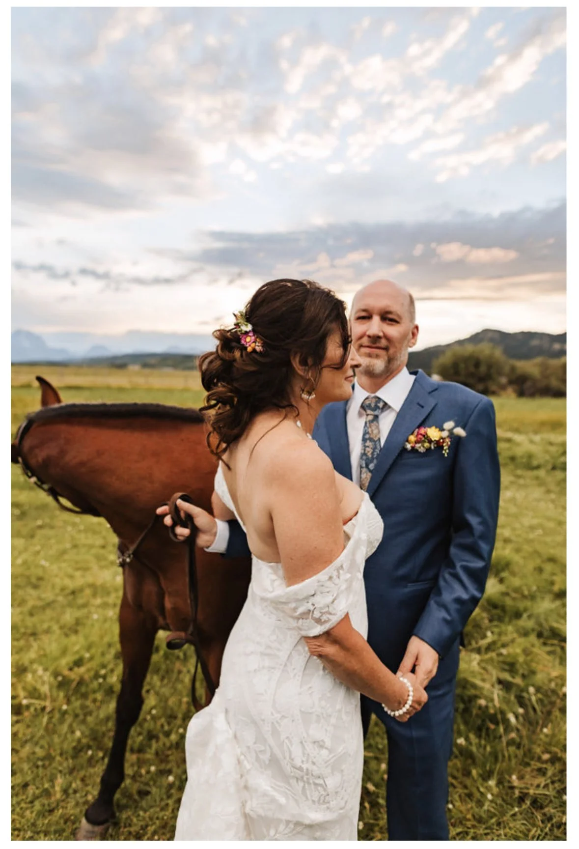 A bride and groom embrace outdoors during their wedding, with a horse in the background, open field, mountains, and a cloudy sky at sunset.