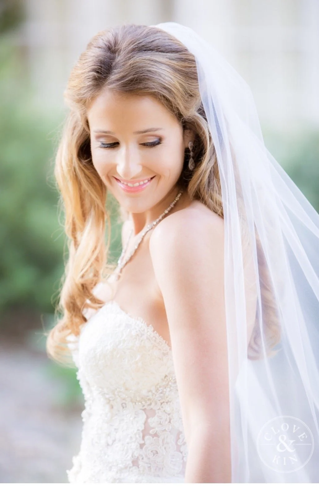 A smiling bride with long, wavy hair wearing a white lace wedding dress and a veil, outdoors with a blurred green background.