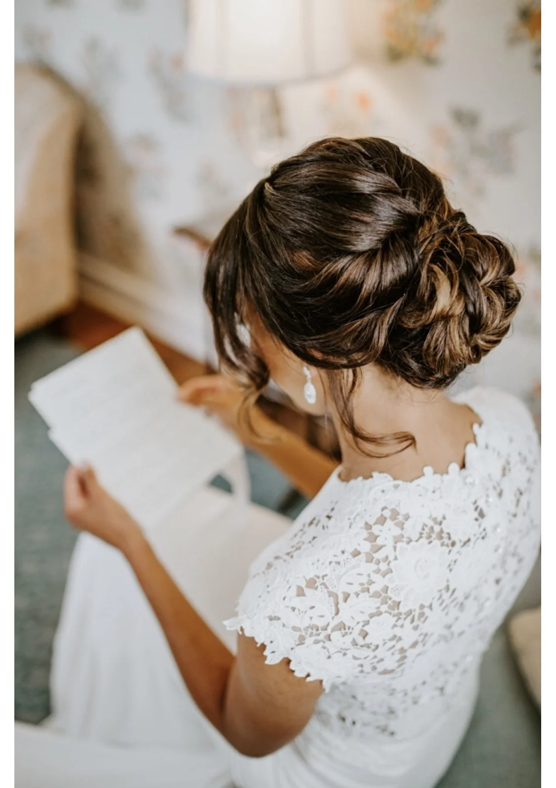 A woman with styled dark brown hair and earrings, wearing a white lace dress, reading a letter or paper while sitting in a room with floral wallpaper.