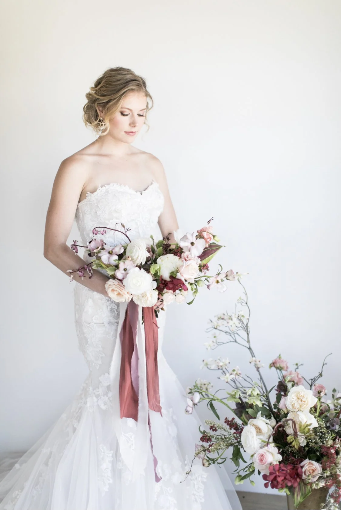 Bride in white lace wedding dress holding a large bouquet of pink, white, and purple flowers with greenery, standing next to a floral arrangement.