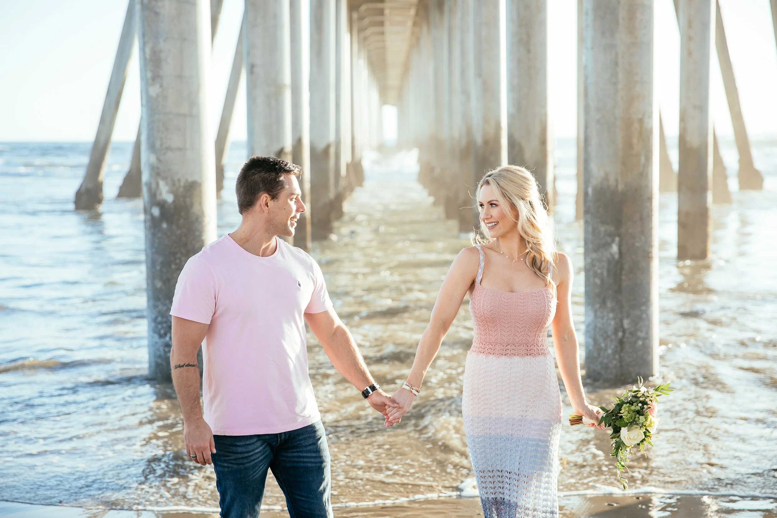 A couple holding hands and walking on the beach under a pier, with the woman holding a bouquet of flowers and wearing a pink dress, while the man wears a light pink T-shirt and jeans.
