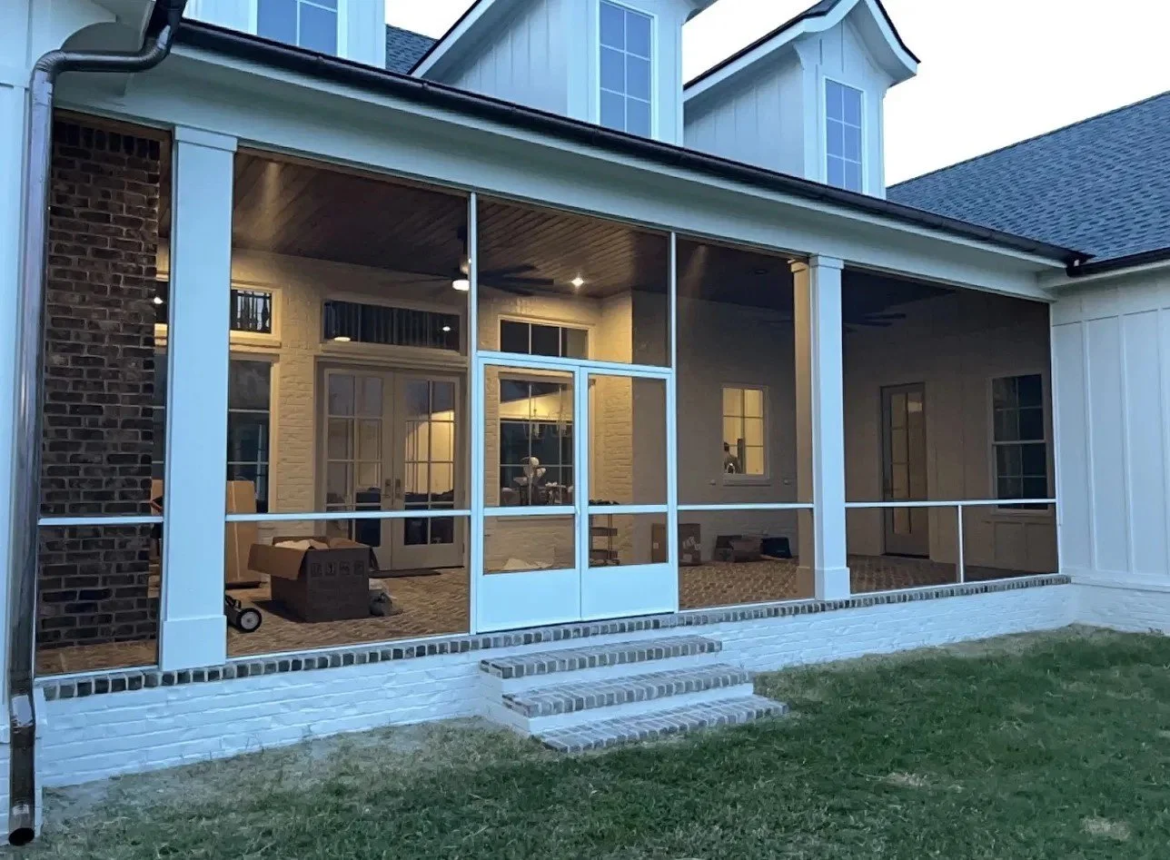 View of a screened porch attached to a house with white walls, brick steps, and potted plants inside.