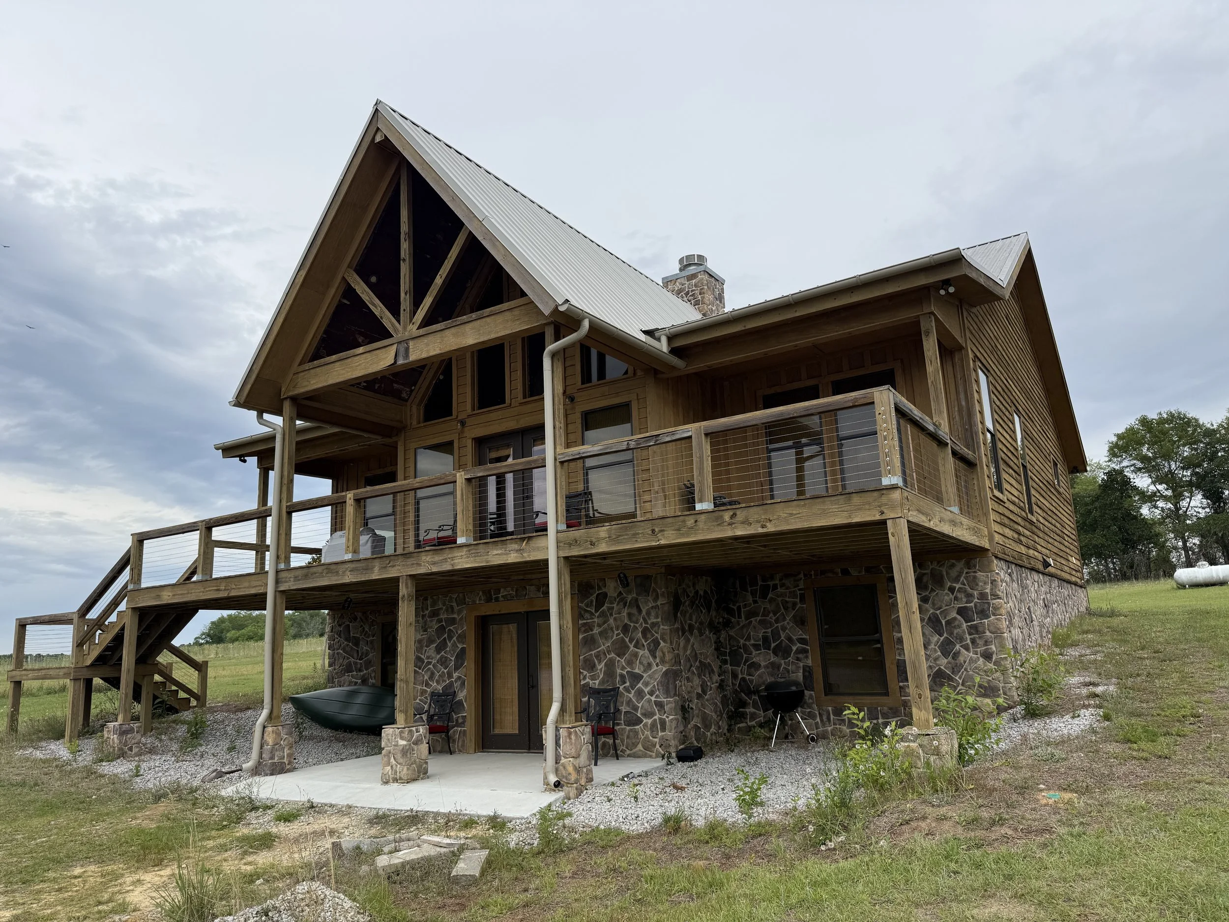 A multi-story house with wooden siding and a stone foundation, featuring large windows, a spacious deck with railings, and an outdoor grill, set in a rural area with grass and trees.