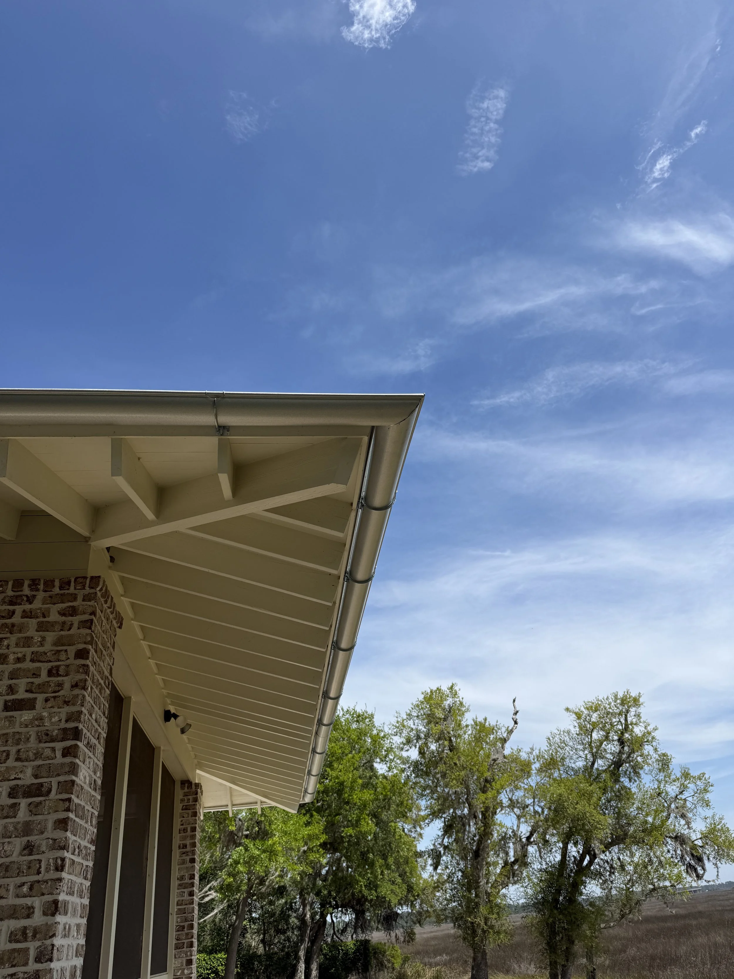 A house with a brick wall, beige siding, and a metal gutter, with a clear blue sky, some white clouds, and green trees in the background.