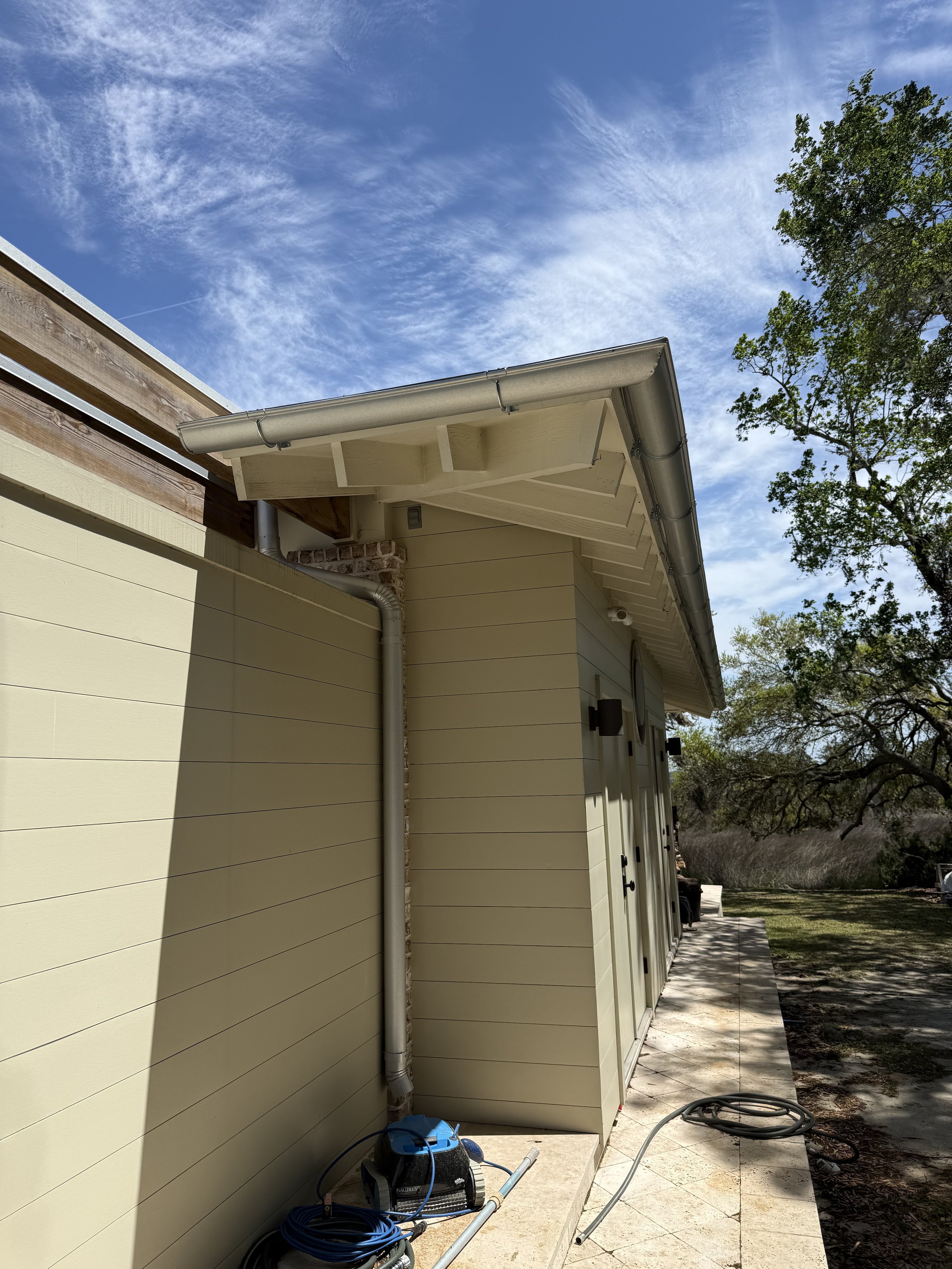 Backyard with house siding, patio, garden hose, and gardening equipment next to the house, with trees and a blue sky with wispy clouds in the background.