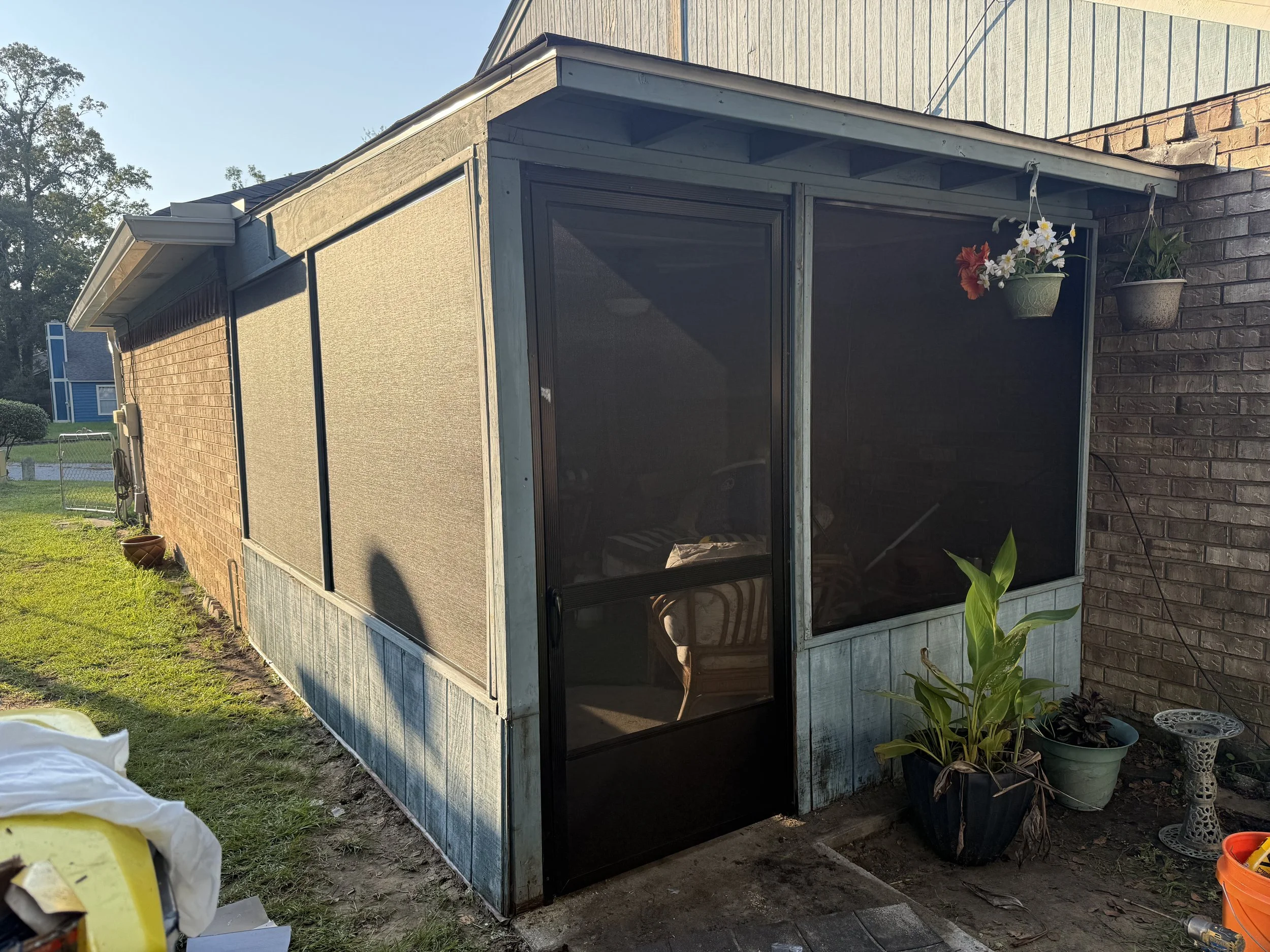 A screened porch attached to a brick house, with hanging flower pots and potted plants outside, and a garden nearby.
