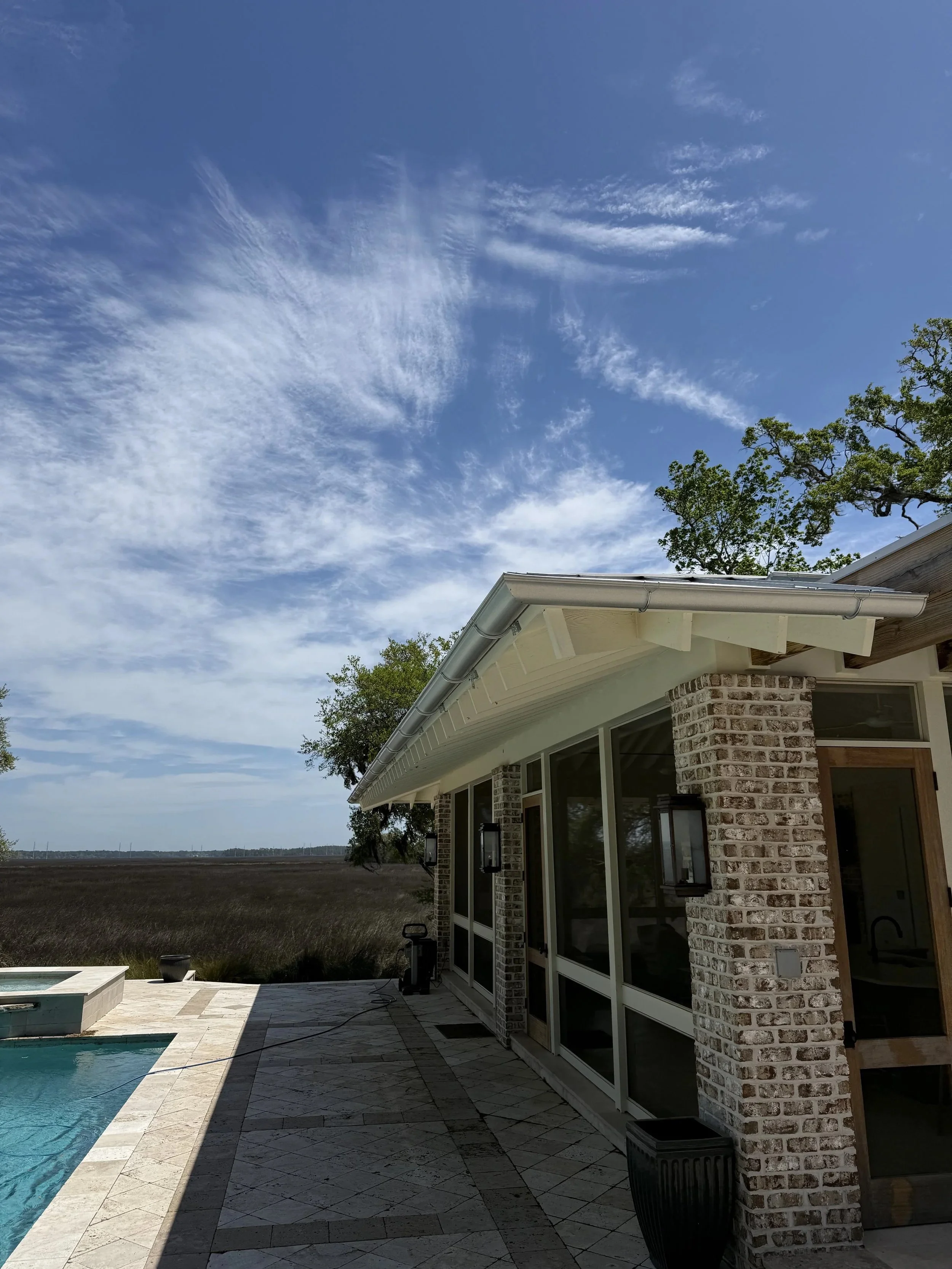 Exterior view of a house with brick walls, large windows, and a metal roof, with a backyard pool area and open field in the background under a blue sky with clouds.
