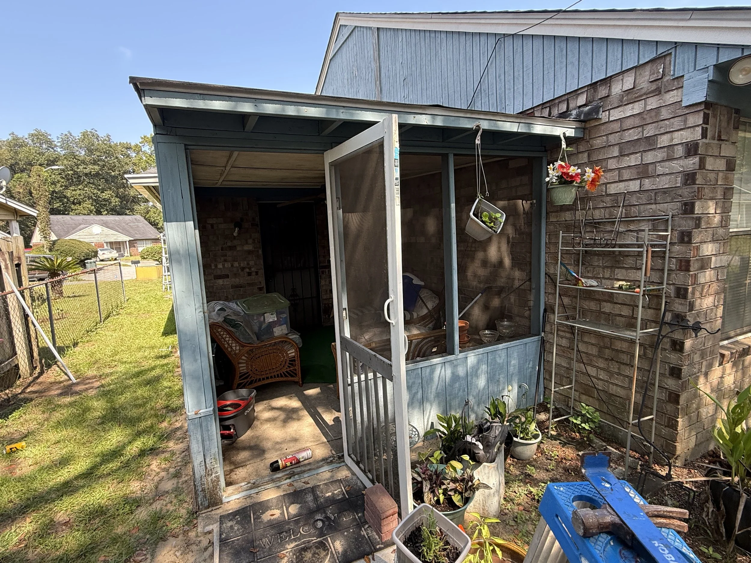 View of a backyard area with a small open porch attached to a brick house. The porch has a metal roof and some hanging flower baskets. The backyard is fenced, with some outdoor furniture, gardening tools, and various potted plants visible. The scene 