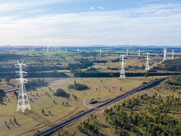 A vast landscape of grassy fields with numerous electrical transmission towers and power lines running through it. In the background, more towers extend toward the horizon under a partly cloudy sky.