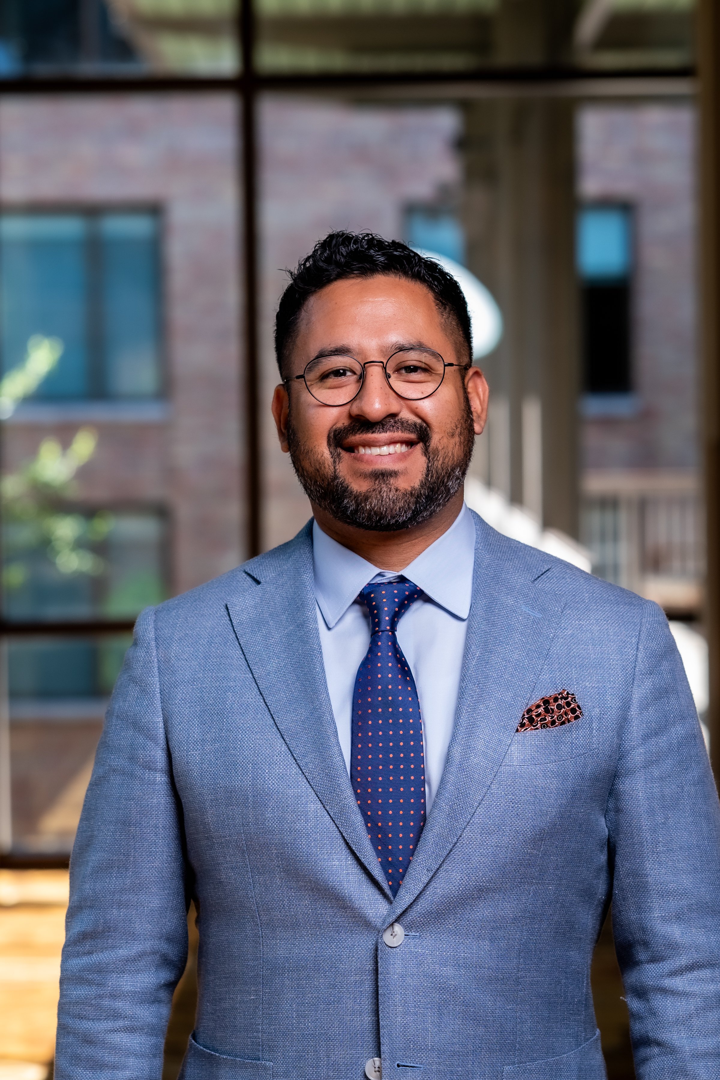 Smiling man in a light blue suit, white shirt, blue tie with red dots, and pocket square, standing outdoors with a building and windows in the background.