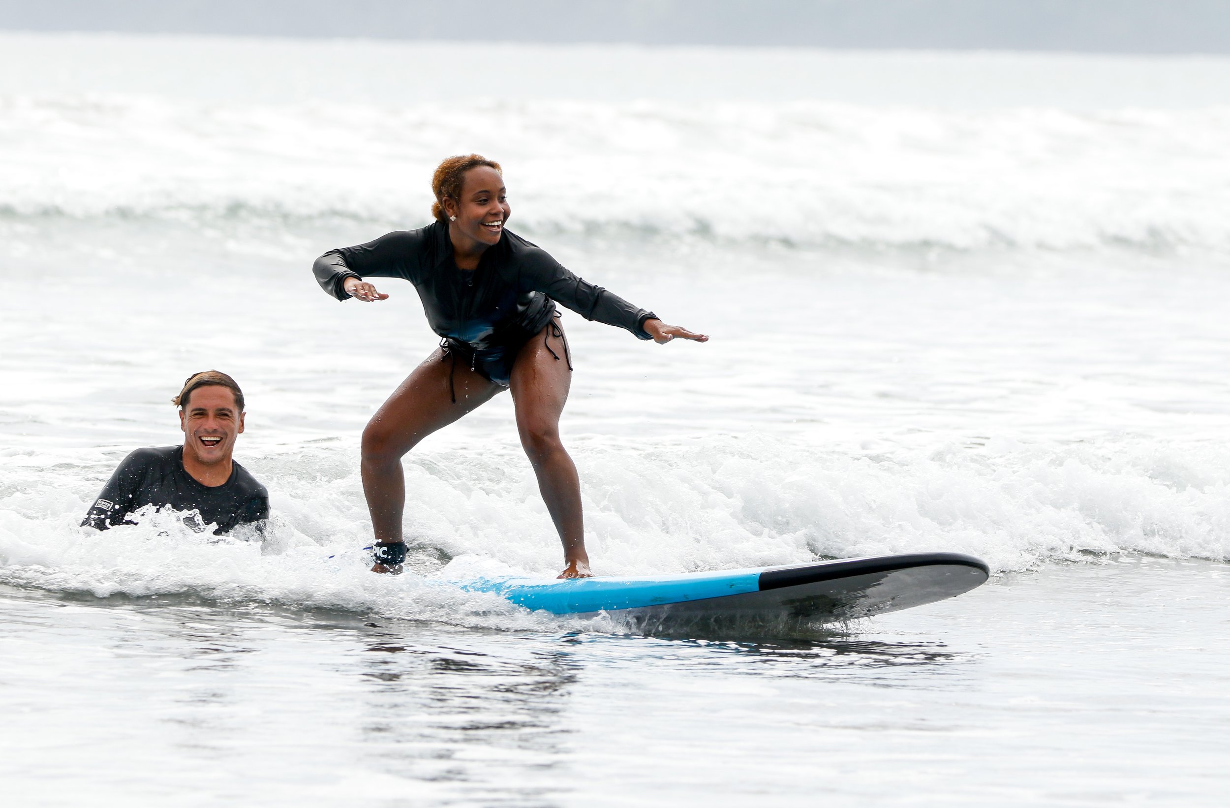 Two people riding a surfboard in the ocean, with waves and foam around them, smiling and enjoying the ride.