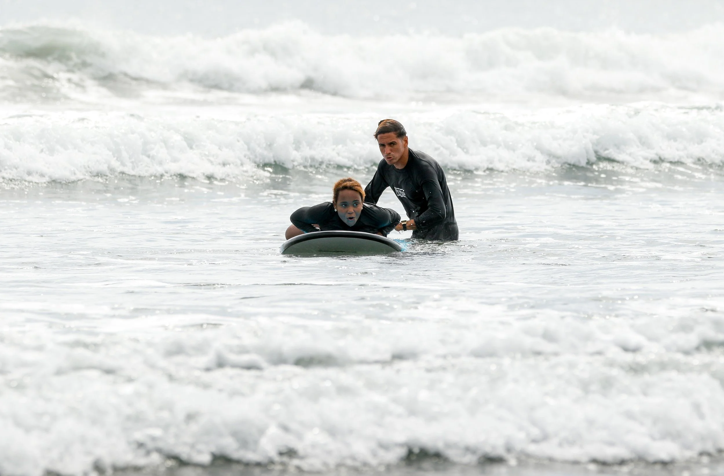 A woman with short curly hair in a black wetsuit lying on a surfboard in the ocean with a man in a black wetsuit helping her, and ocean waves in the background.